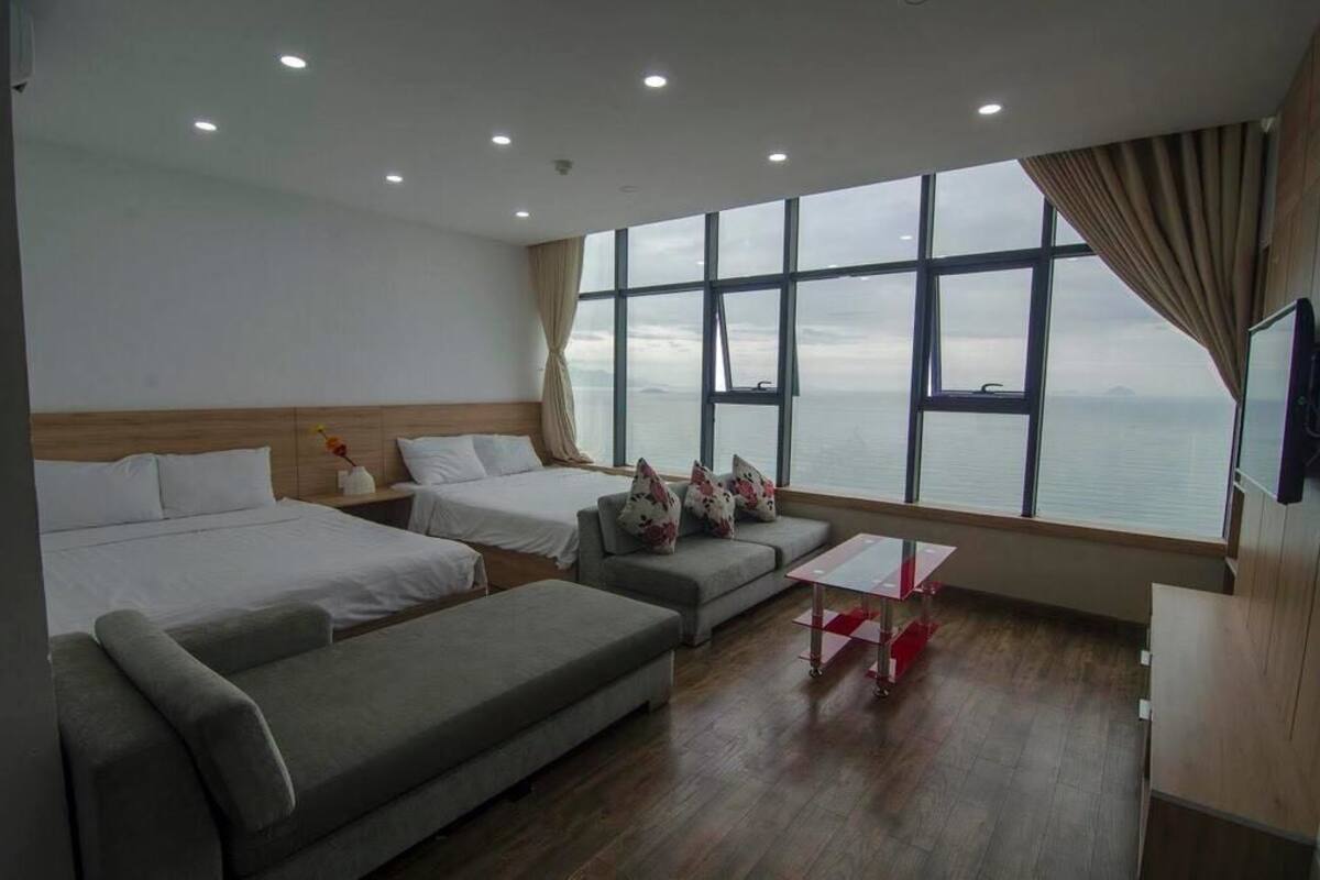 A hotel room with two beds, a sofa, a small table, and a large window offering a view of the sea.