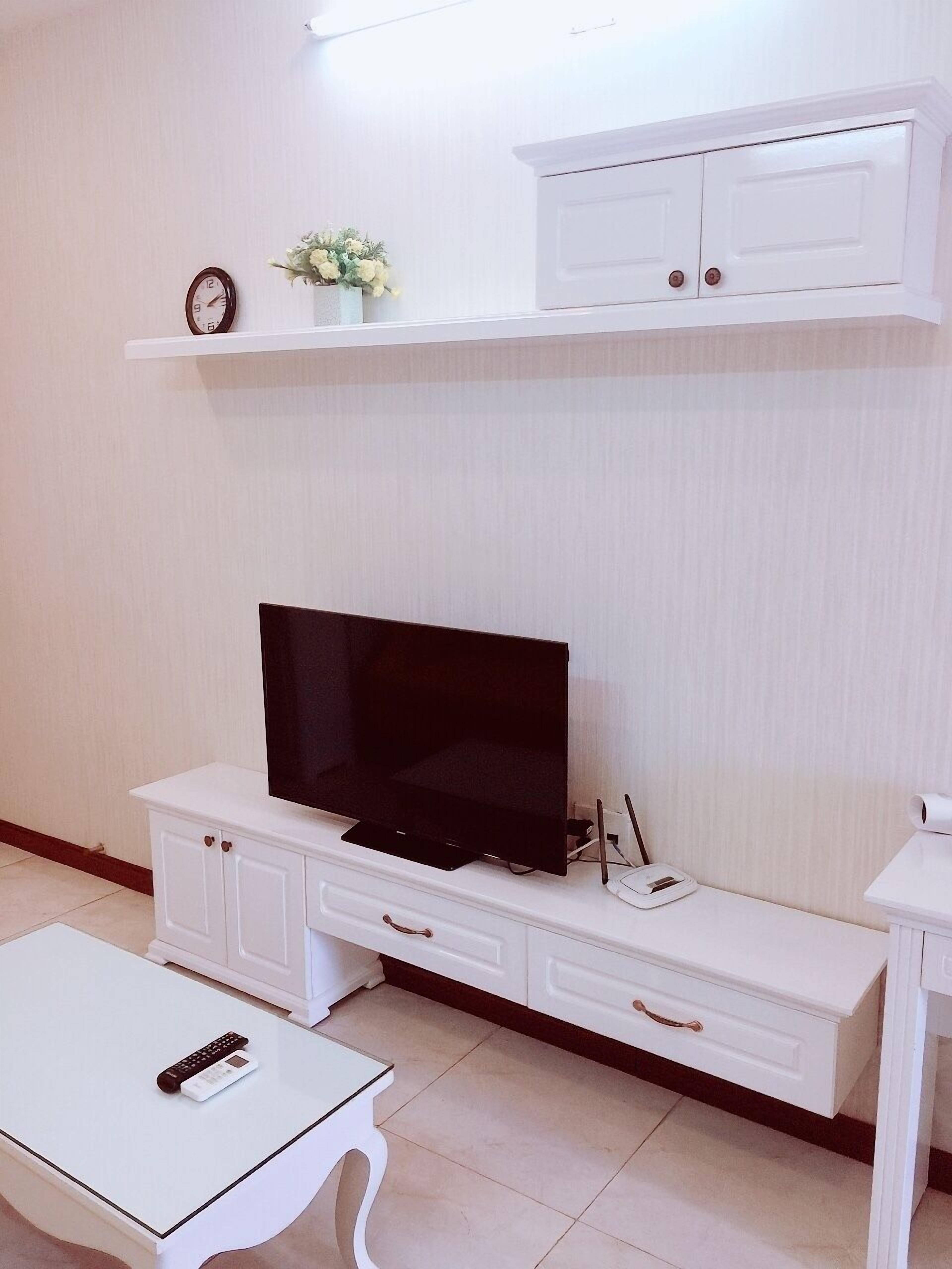 A living room with a flat-screen TV on a white entertainment unit, a small table with a remote control, and a shelf with a clock and flowers.