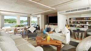 A modern living room with a large TV, comfortable seating, a bookshelf, and a view of the outdoors. - AYANA Lako di'a (Labuan Bajo)