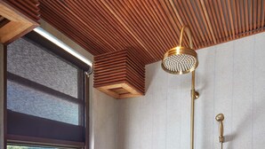 A bathroom with a wooden slatted ceiling, a shower with a gold showerhead, a bench, and a window overlooking a body of water. - AYANA Lako di'a (Labuan Bajo)