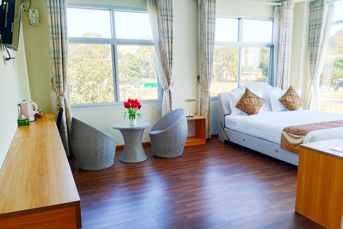 A hotel room with a wooden floor, a bed with white bedding and patterned pillows, a small round table with a vase of red flowers, two wicker chairs, and a bedside table.