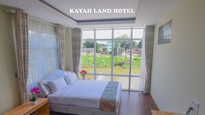 Quarto de hotel com uma cama grande, mesinhas de cabeceira de madeira, uma escrivaninha com telefone e vista para áreas verdes e edifícios através de janelas amplas.
