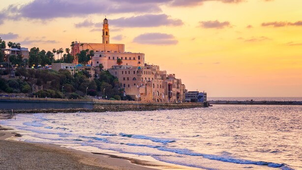 Un paisaje urbano costero con edificios históricos, una torre destacada y una playa con olas.