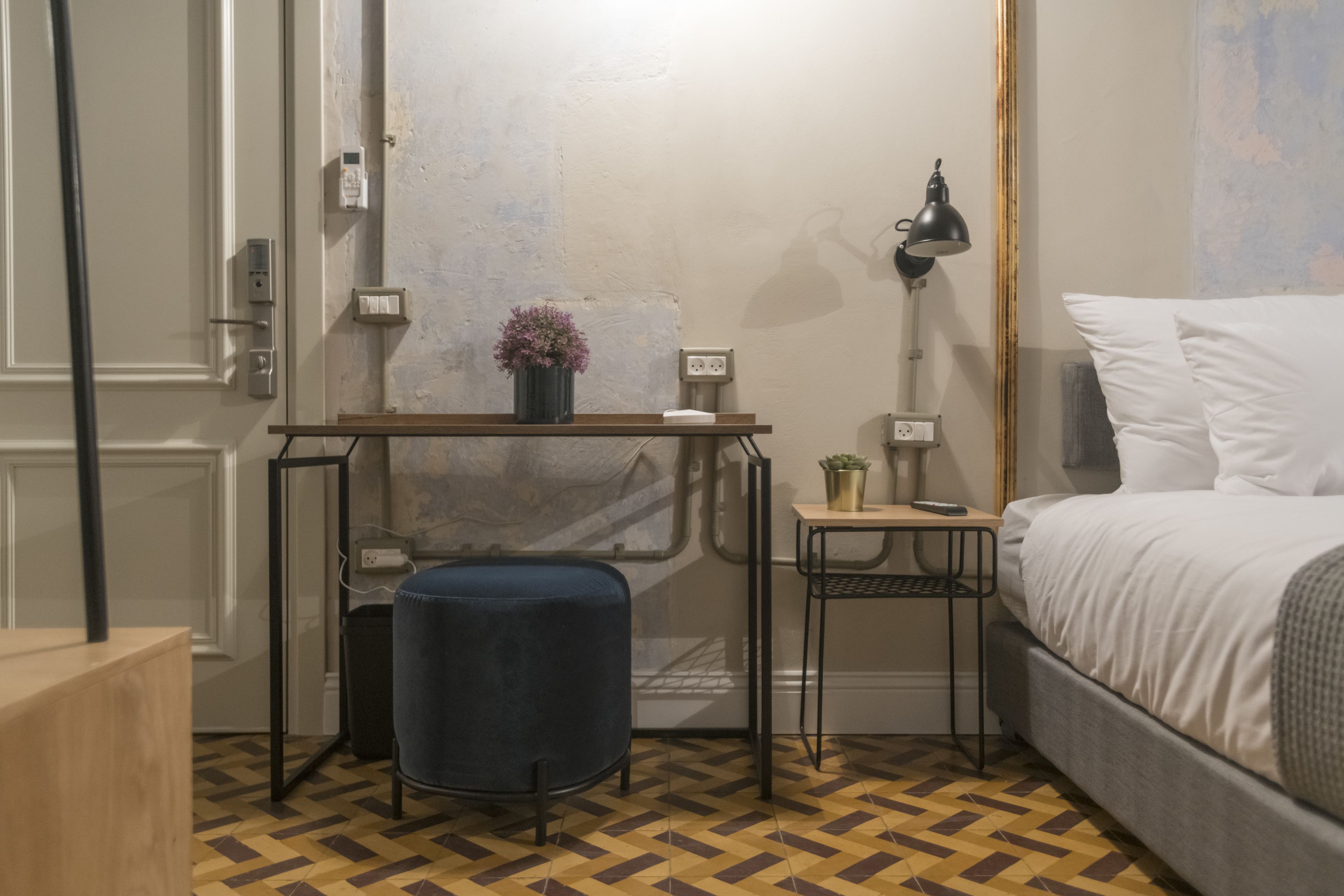 Quarto de hotel pequeno e compacto, com uma cama, escrivaninha com luminária, cadeira, mesa pequena com uma planta e um banquinho azul.