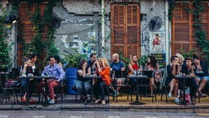 Un café al aire libre con mesas y sillas, frente a un muro de ladrillo con contraventanas de madera y carteles.