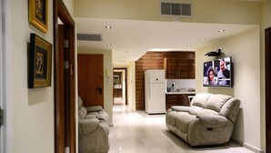 A spacious living area with two beige sofas, a kitchen with a refrigerator, and a television mounted on the wall. - Garden Residence (Eilat)