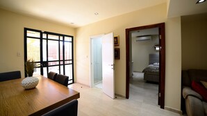 A spacious living area with a wooden dining table, a sofa, and a doorway leading to a bedroom with a bed and an air conditioning unit. - Garden Residence (Eilat)