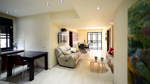 A modern living room with a sofa, dining table, and chairs. There is a kitchen area with a refrigerator and a window. - Garden Residence (Eilat)