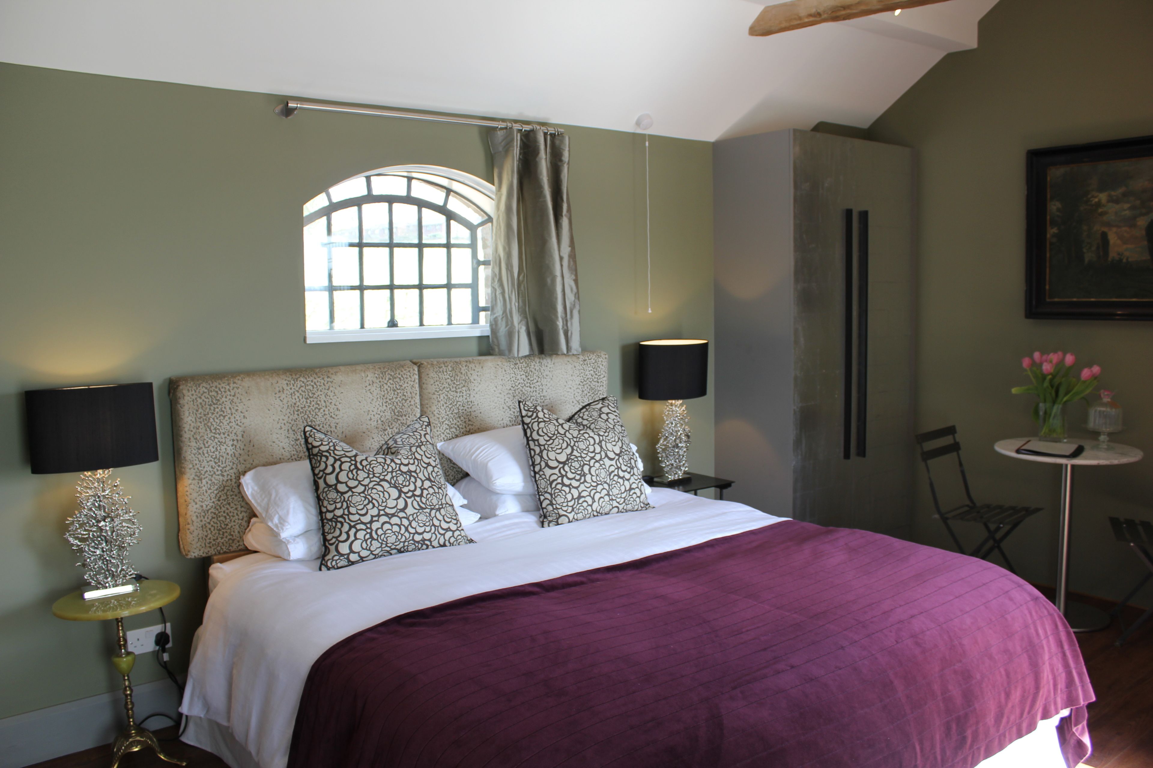 A bedroom with a bed, two bedside lamps, a window with curtains, a small table with a vase, and a chair.
