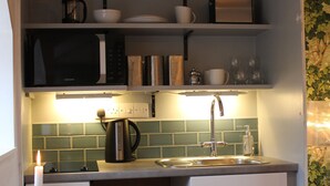 Een kleine keuken met een magnetron, waterkoker en verschillende serviesdelen op planken. Een eettafel met borden en een kaars is te zien.