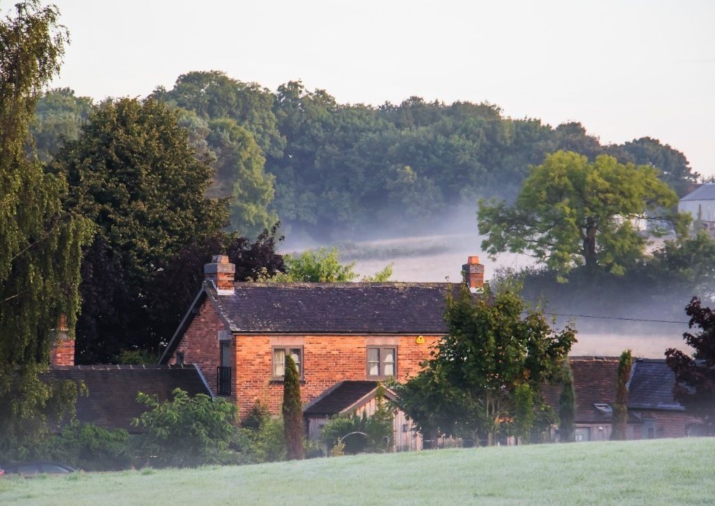 Une maison de campagne aux murs en briques, entourée de verdure et d’arbres.