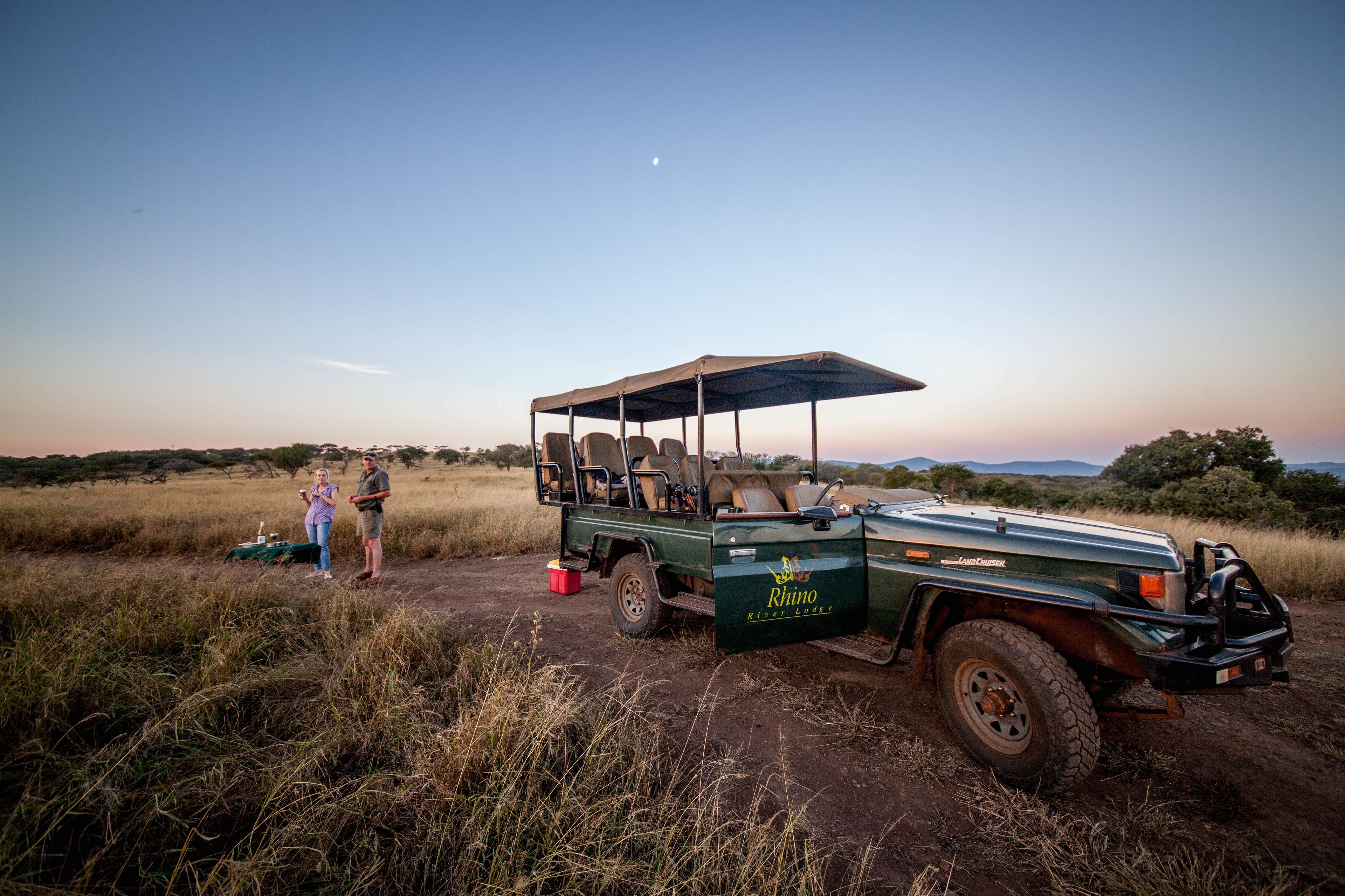 A safari vehicle with a canopy, a table with a cooler, and two people standing nearby in a grassy field.