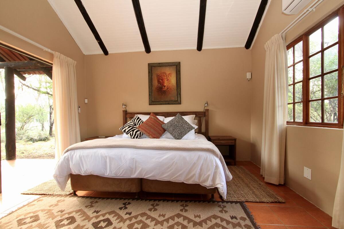 Une chambre à coucher avec un grand lit, une tête de lit en bois, une table de chevet et une fenêtre avec des rideaux.