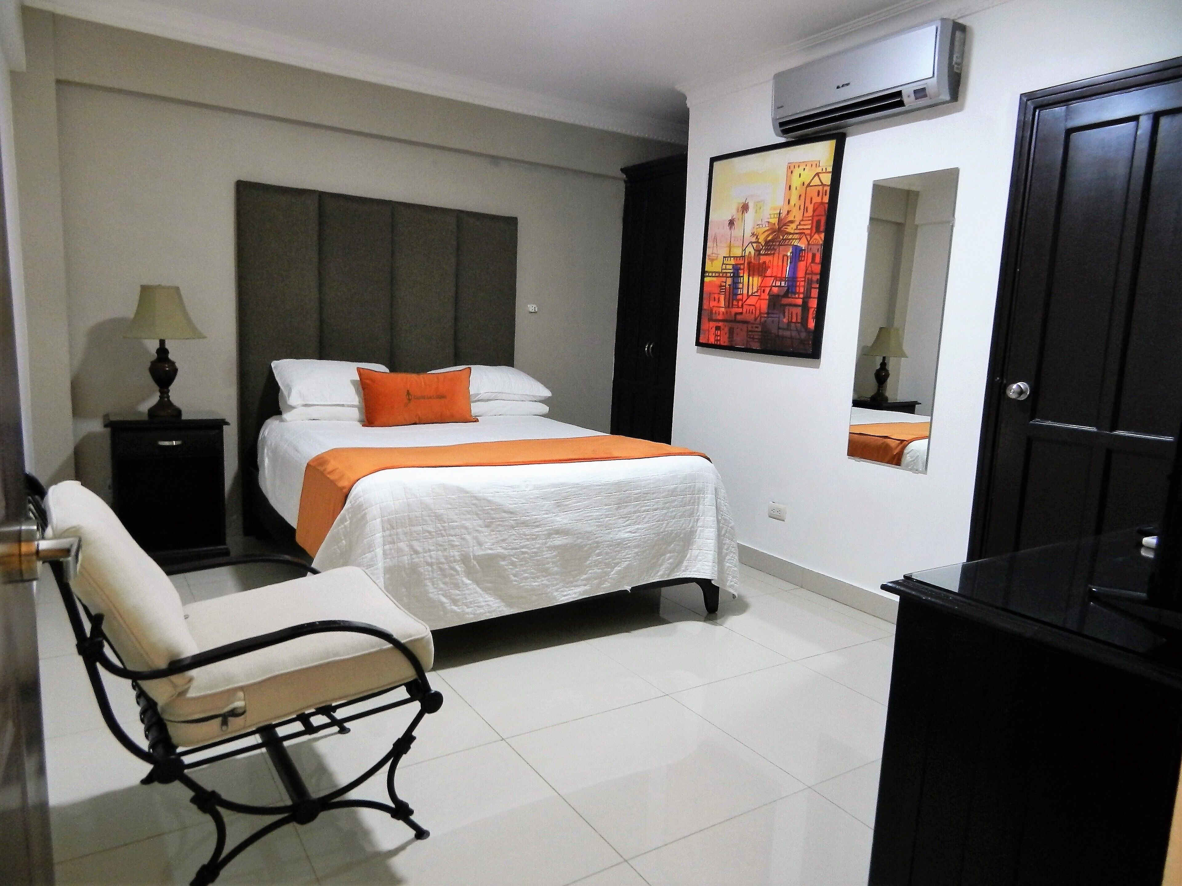 A hotel room with a bed, a chair, a nightstand, an air conditioner, a painting, and a door.