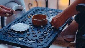 A close-up of a black metal table with a white plate and a brown bowl, surrounded by people holding cups.