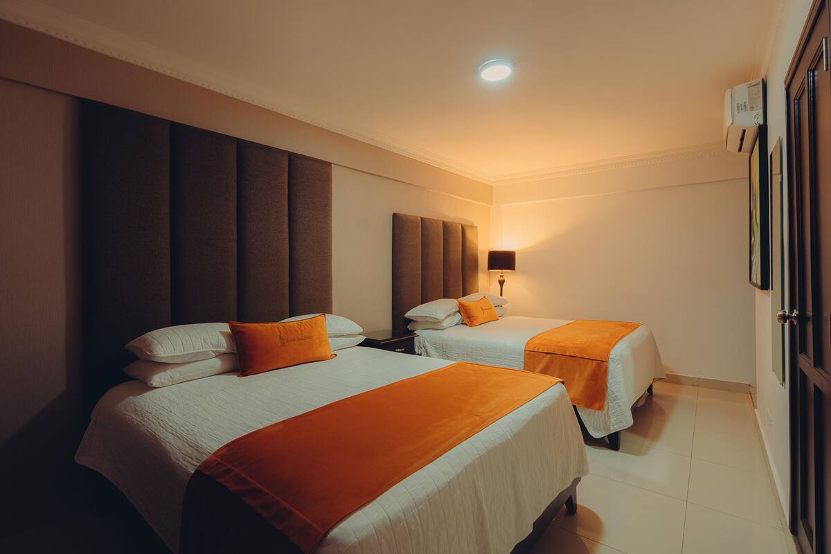 A hotel room with two beds, a headboard, a nightstand, a lamp, and a wall-mounted air conditioner.