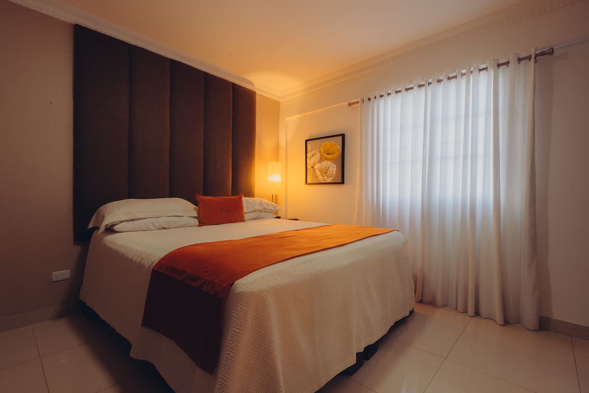 A neatly made bed with a white and orange bedspread, a single orange pillow, and a wall-mounted lamp.