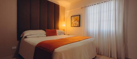A neatly made bed with a white and orange bedspread, a single orange pillow, and a wall-mounted lamp.