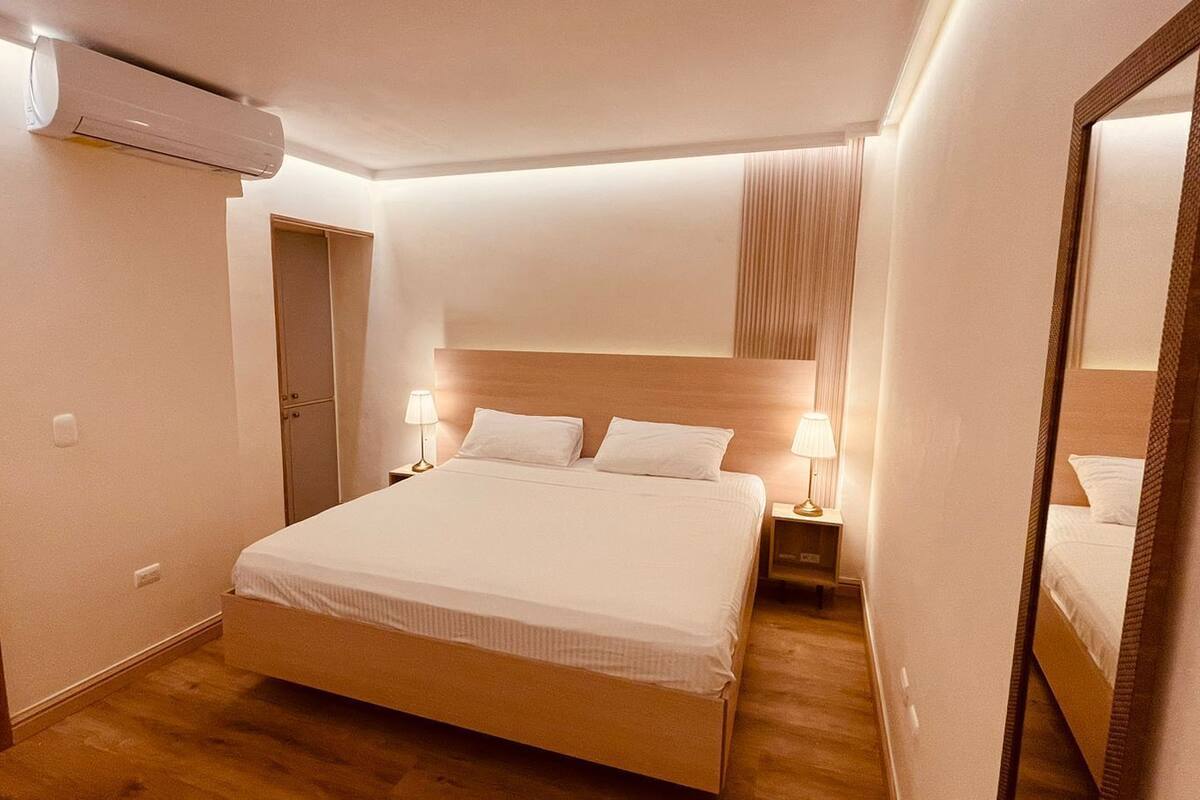 A hotel room with a bed, bedside lamps, a mirror, and an air conditioning unit.