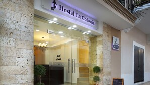 A hotel entrance with a sign reading "Hostal La Colonia," featuring a modern glass facade, a staircase leading inside, and two potted plants outside.