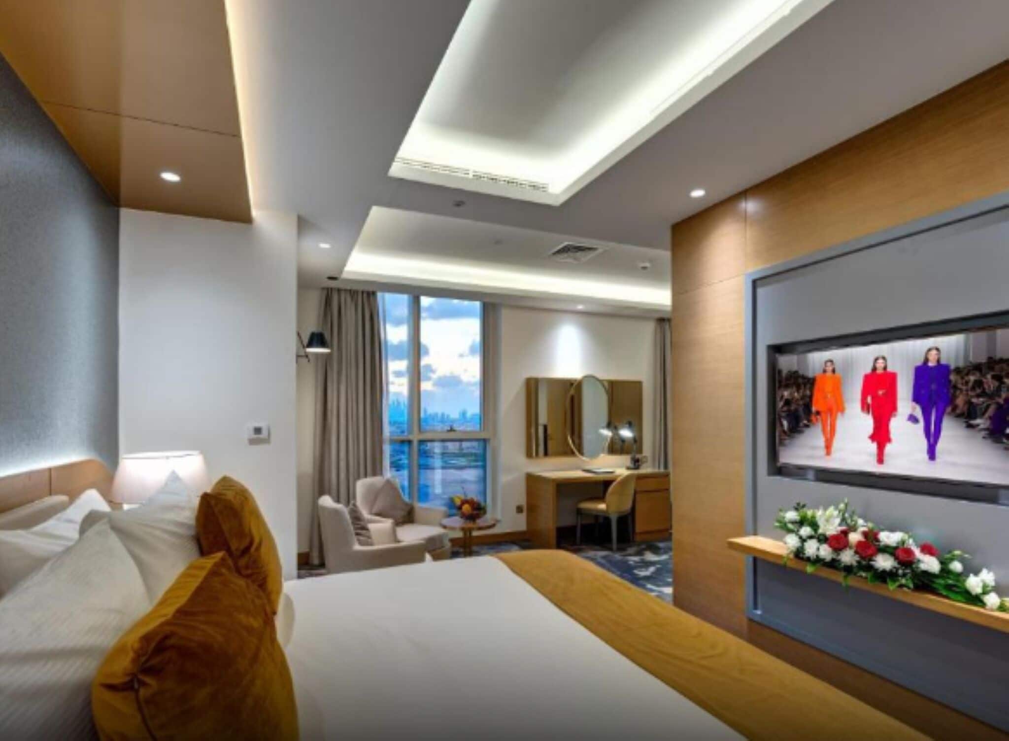 A hotel room with a bed, a desk, a chair, a large window, and a TV screen displaying a fashion show.