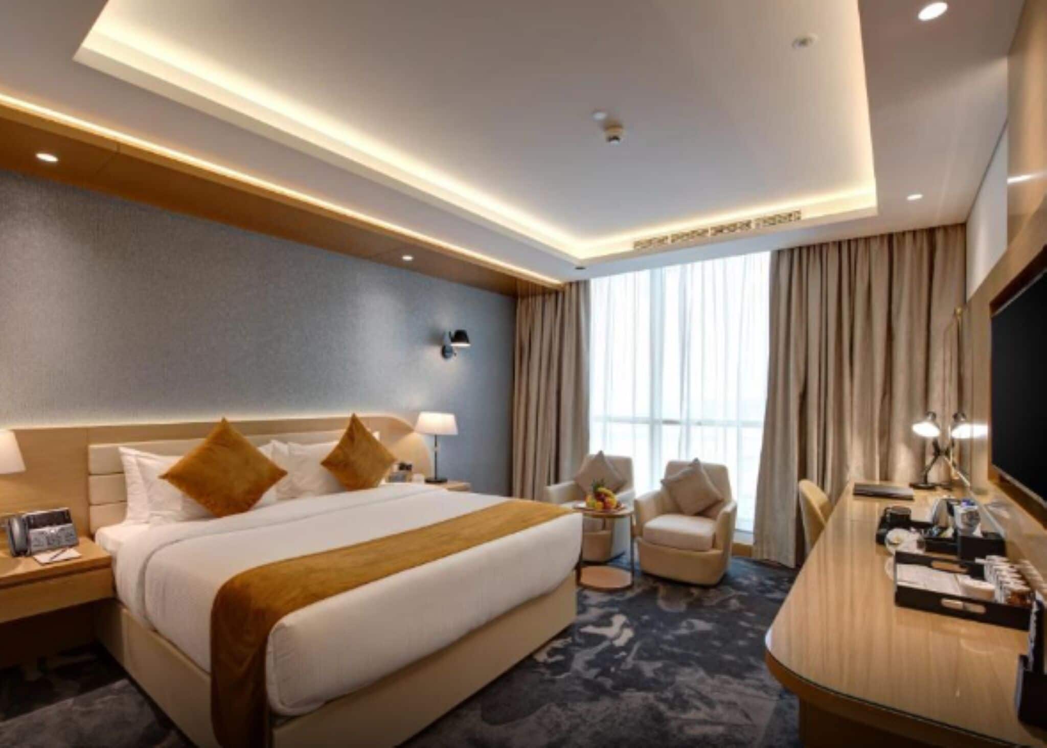 A hotel room with a bed, bedside tables, a desk, a chair, and a large window with curtains.