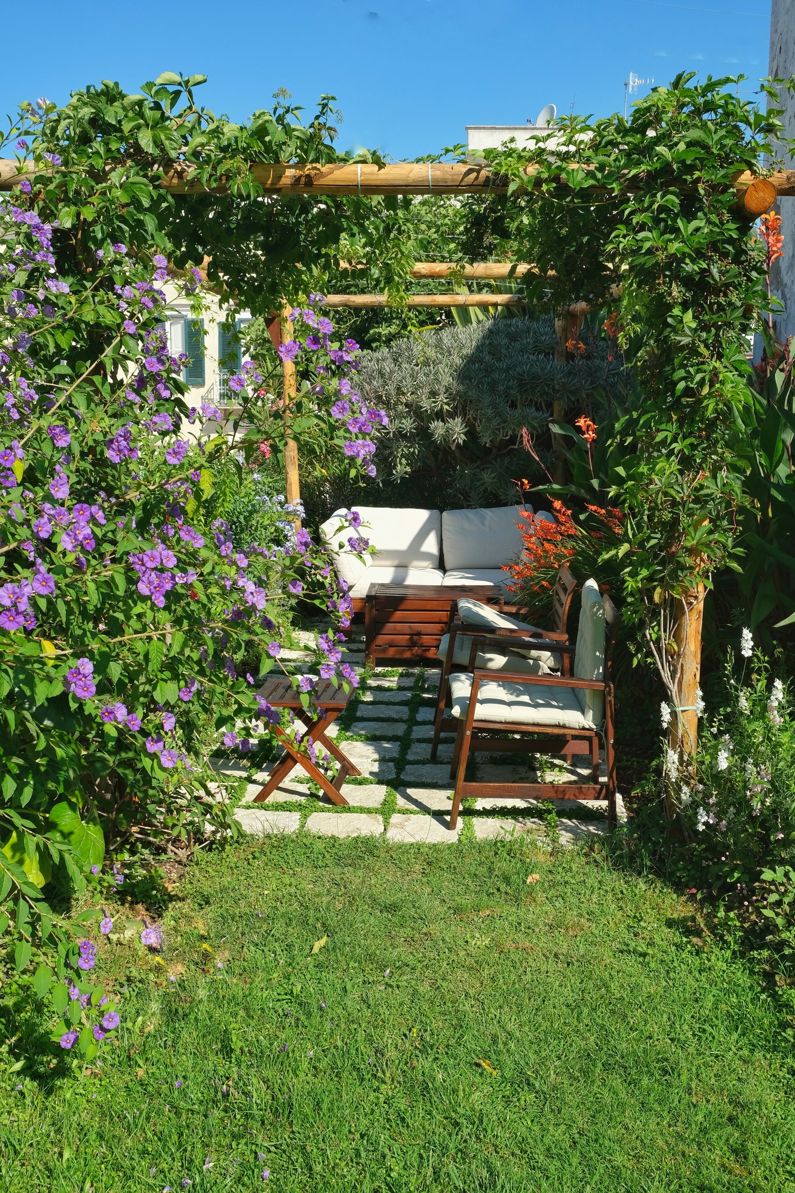 A garden with a wooden pergola, a white outdoor sofa, and a wooden chair.