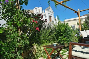 A garden with a white building in the background, a wooden table with a bowl, and a white sofa. - Le Cantinelle (Anacapri)