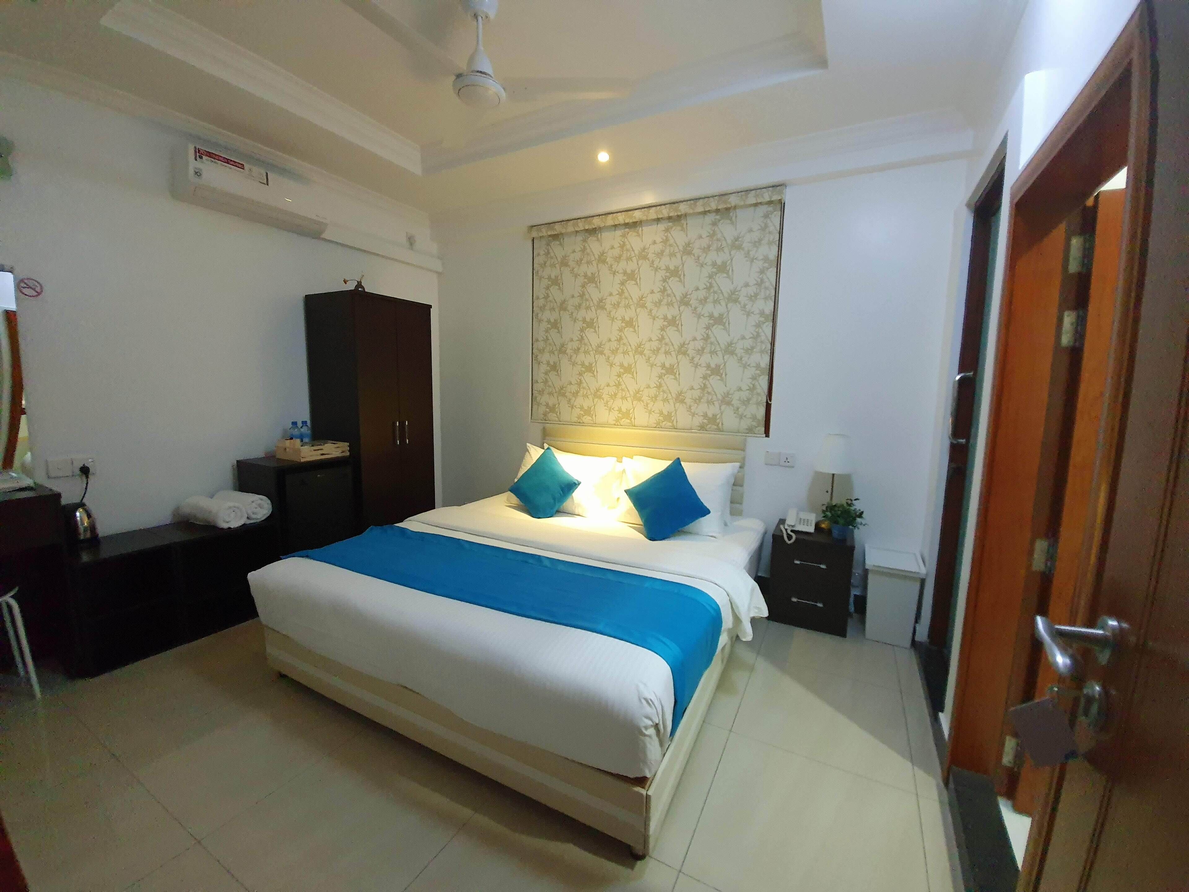 A hotel room with a large bed, a wardrobe, a bedside table, a nightstand, and a window with a patterned curtain.