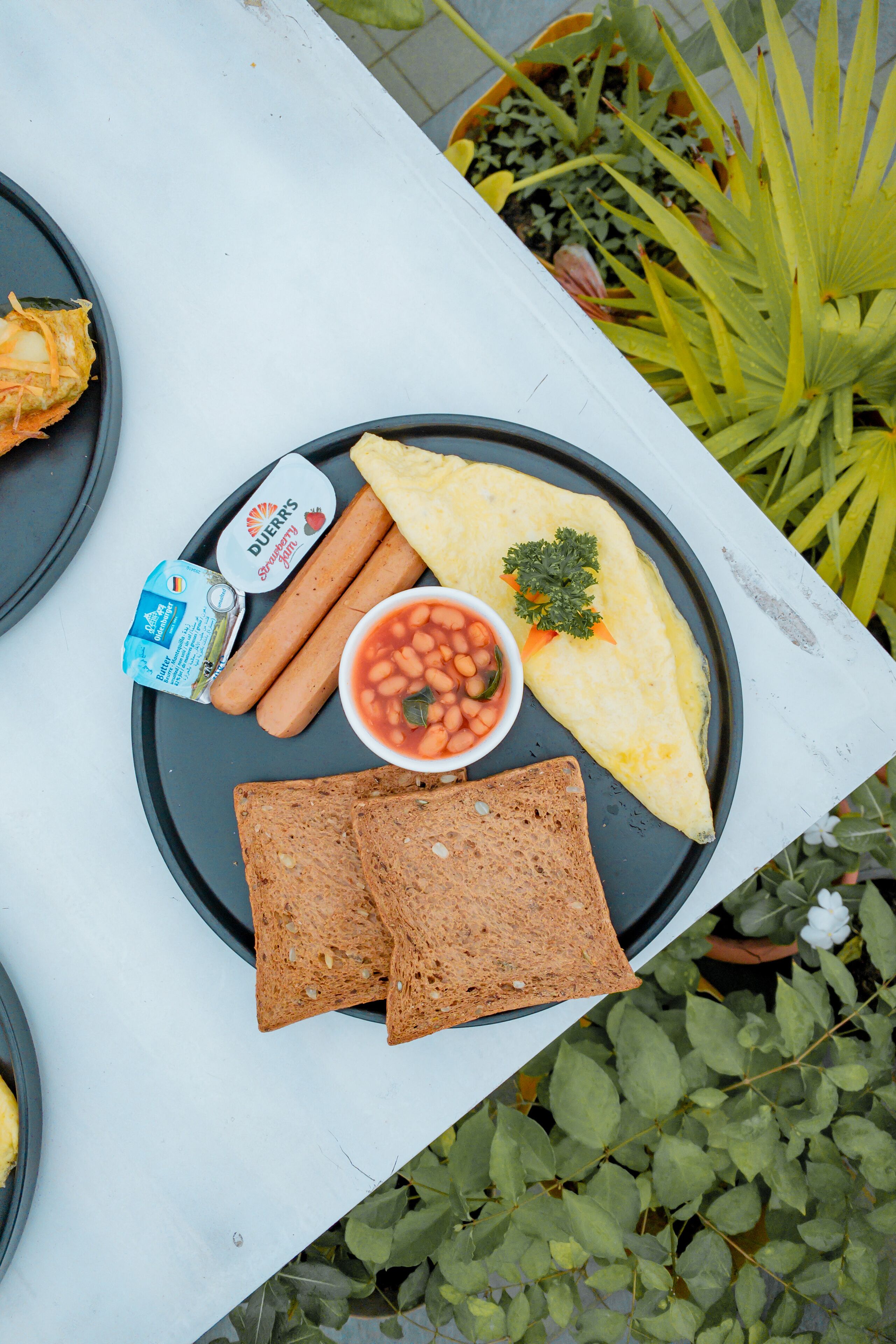 A meal with a plate containing hot dogs, beans, toast, and an omelet, with a butter container and a drink.