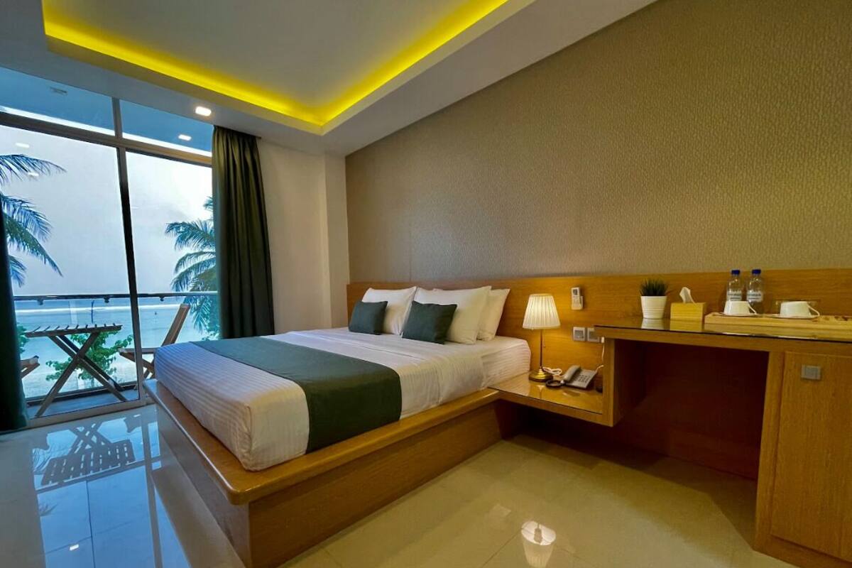 Hotelkamer met een bed, nachtkastje, lamp en uitzicht op palmbomen door grote ramen.