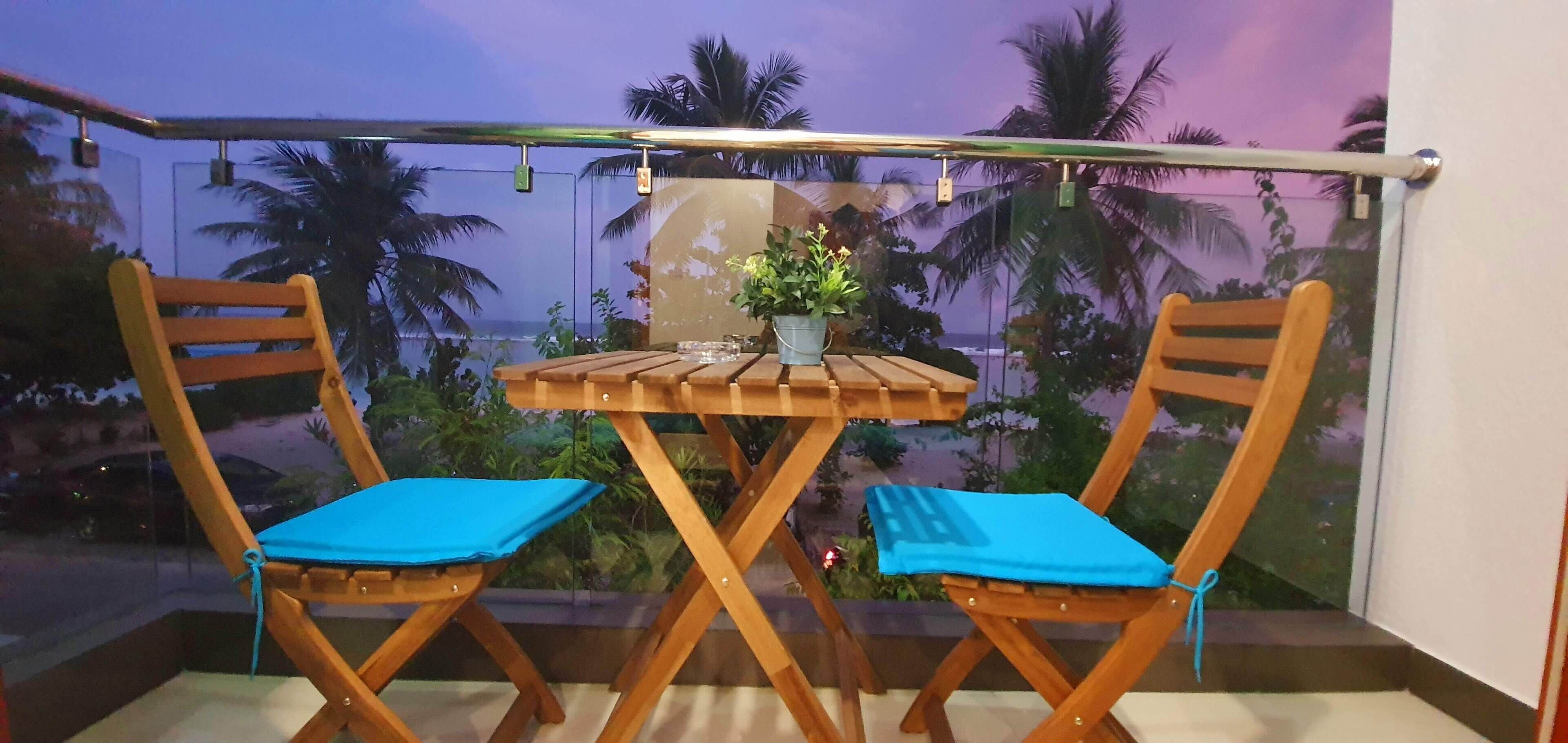 A balcony with a wooden table and two chairs, a potted plant, and a view of palm trees and the sea.