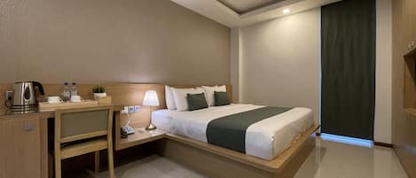 Habitación de hotel con cama, mesita de noche, lámpara y un escritorio pequeño con hervidor y botellas de agua.