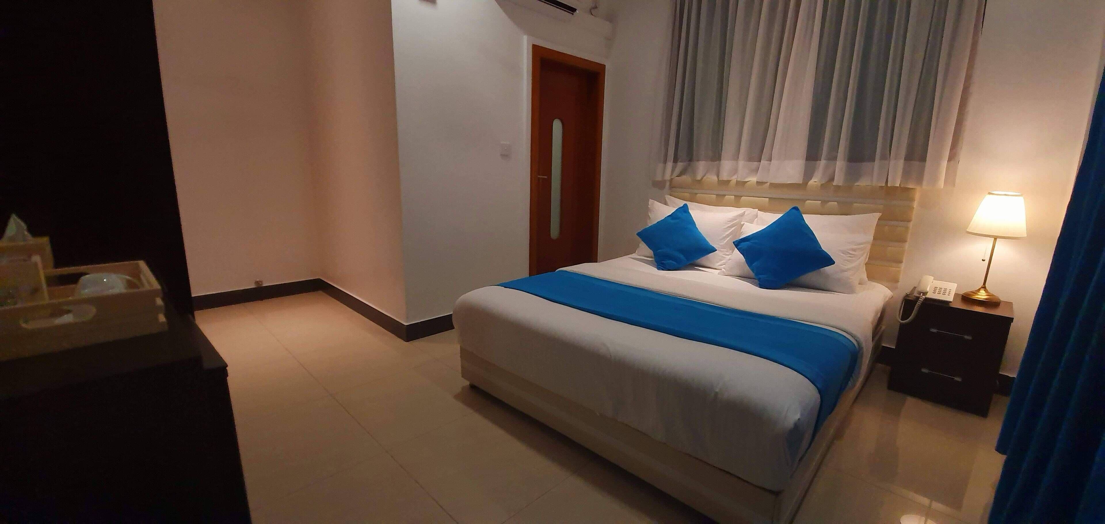 A hotel room with a bed, a bedside table with a lamp, a nightstand, a wooden door, and a window with curtains.