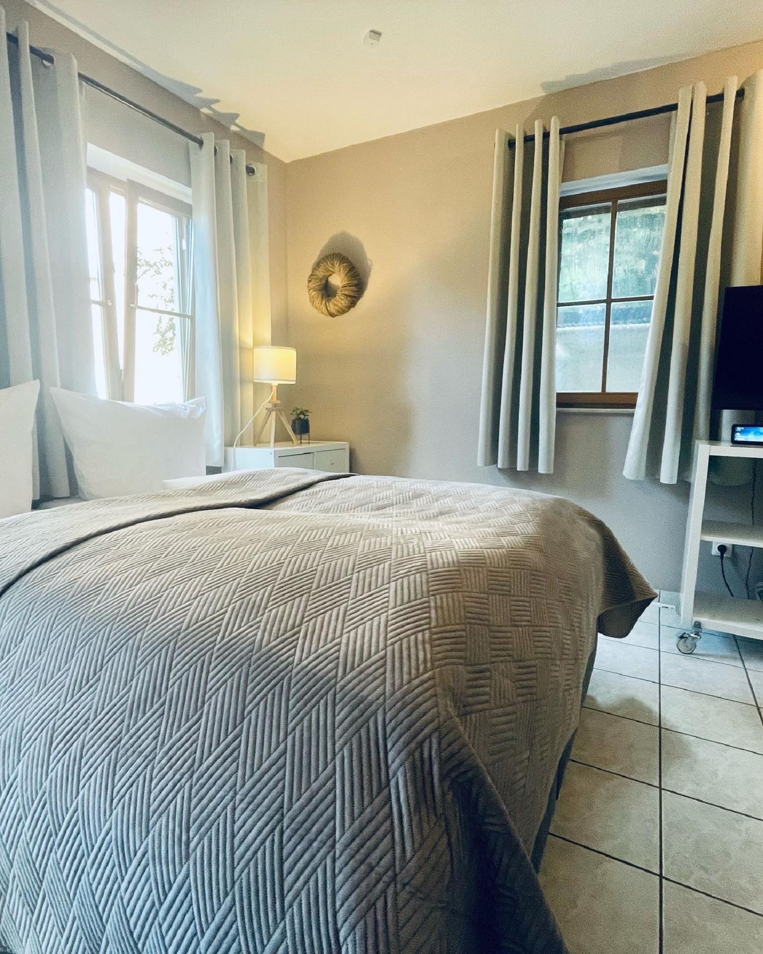 Ein Schlafzimmer mit einem Bett, einem Nachttisch mit Lampe, einem Fernseher auf einem Schrank und Vorhängen an den Fenstern.