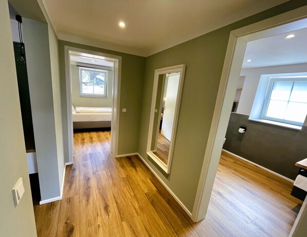 A hallway with wooden flooring, a bed in the background, and a window on the right.