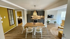A modern apartment with a wooden dining table, white chairs, a kitchen with grey cabinets, and a TV mounted on the wall. - Ferienhof Eichenberg (Haundorf)