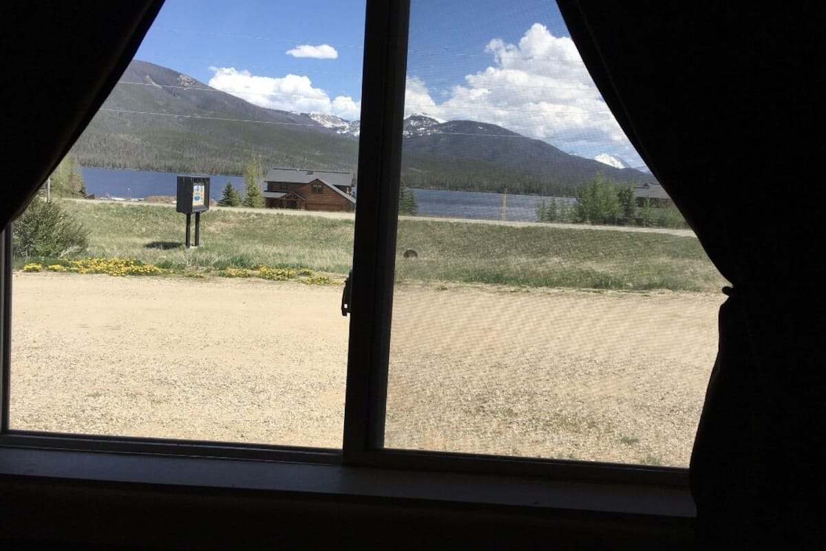 A view from inside a room with a large window overlooking a lake, mountains, and a wooden cabin.