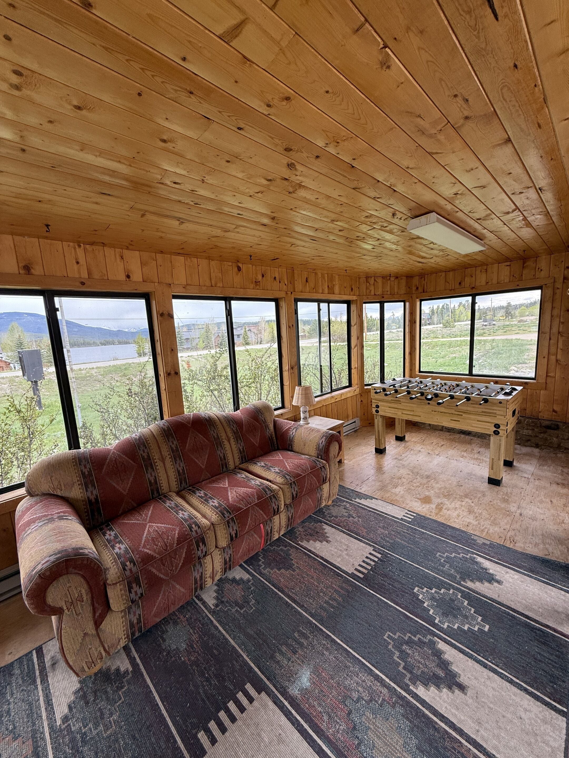 A cozy room with a wooden ceiling, large windows, a sofa, and a foosball table.
