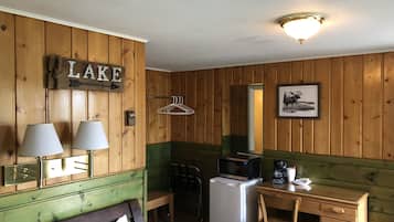 A room with wooden walls, a bed with a green headboard, a desk with a chair, a refrigerator, a microwave, and a framed picture on the wall.
