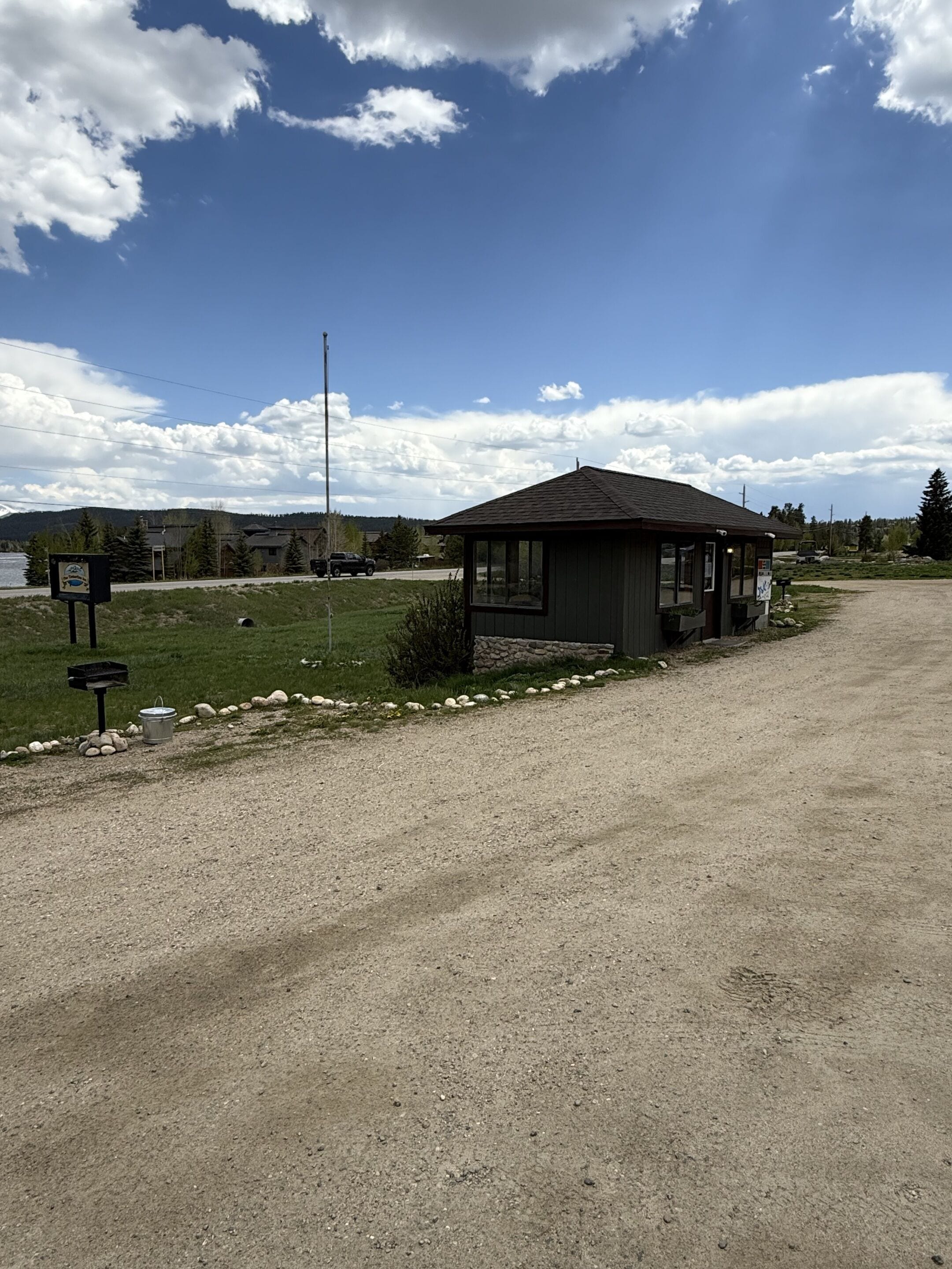 A small, single-story building with a sloped roof, likely a cabin or a small house, situated on a gravel driveway with a grassy area in front.