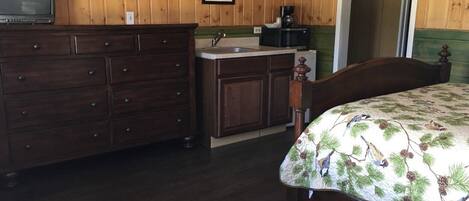 Una habitación con un aparador de madera, una cama con cubrecama de motivos naturales, una pequeña kitchenette y un cuadro en la pared.