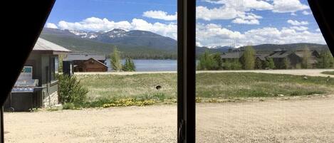 A view through a window of a building with a gravel yard, a wooden cabin, and a lake with mountains in the background.