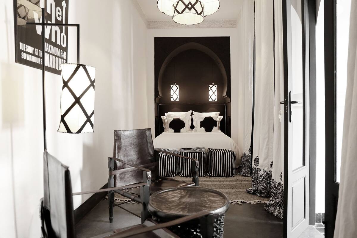 A hallway with a bed, a chair, and a small table. There are patterned cushions on the bed and a decorative lamp on the wall.