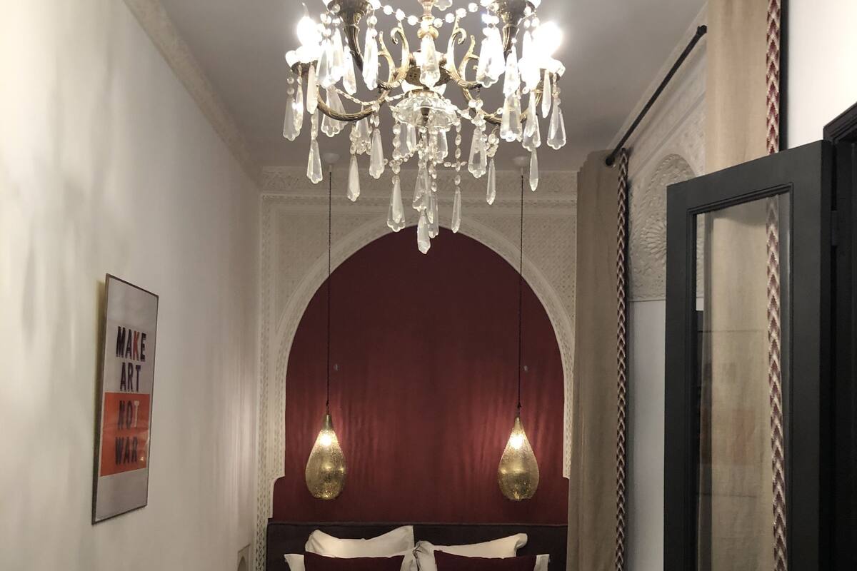 A bedroom with a chandelier, a bed with patterned bedding, a framed poster on the wall, and a door with curtains.