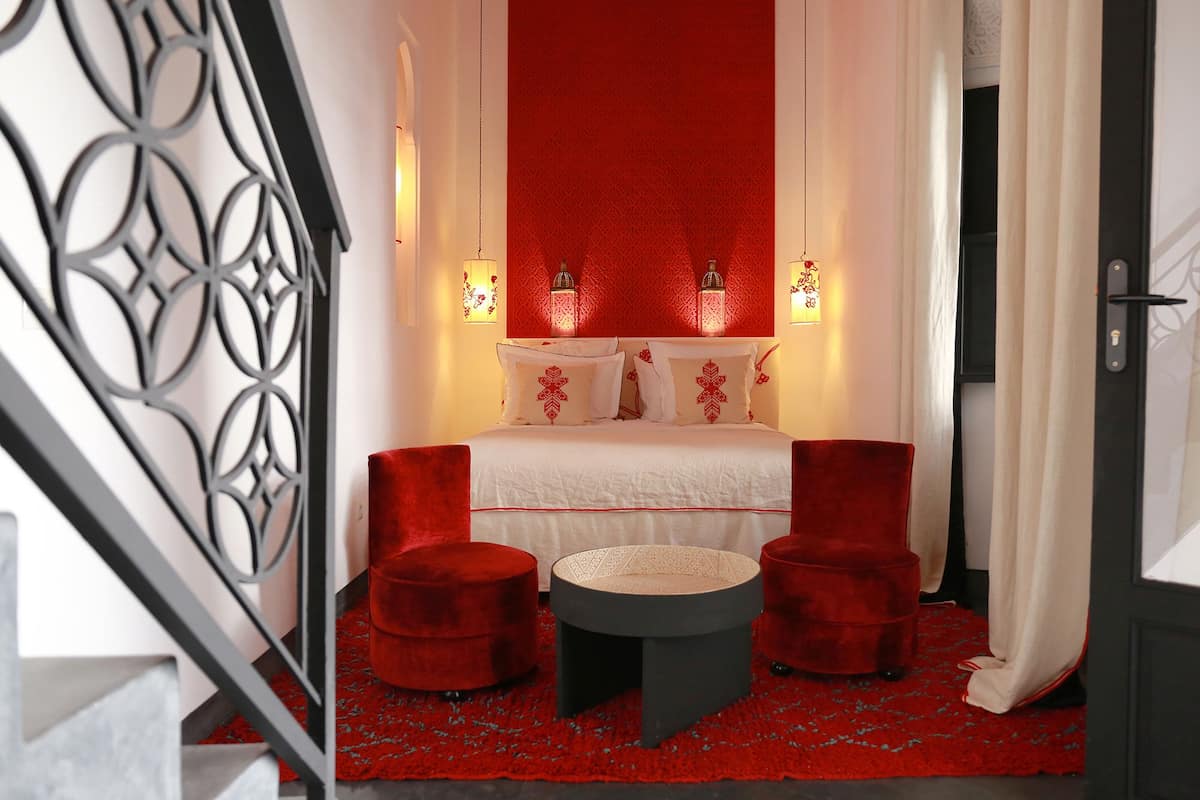 A bedroom with a bed, two red chairs, a small table, and two wall-mounted lamps.