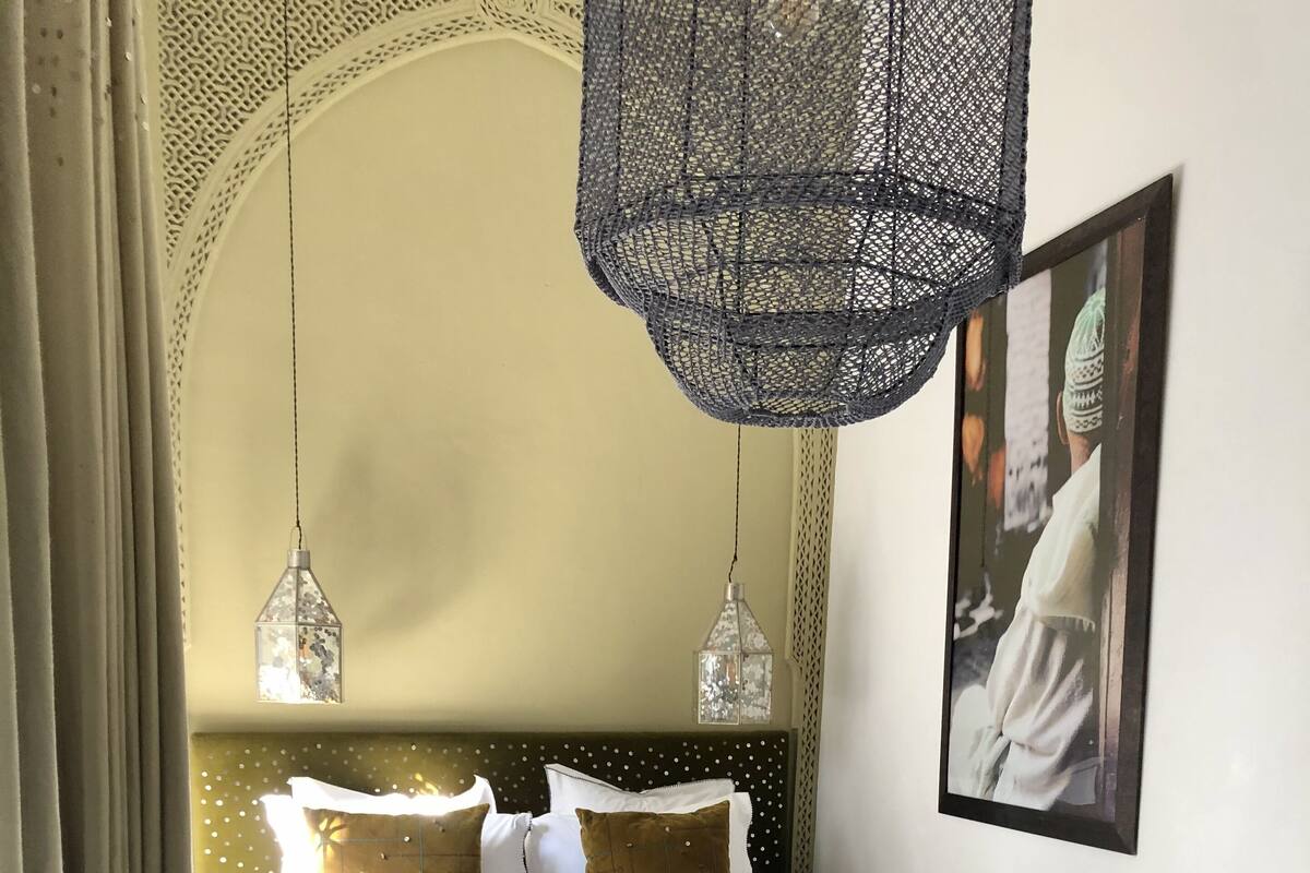 A neatly made bed with a patterned bedspread and pillows, a bench at the foot of the bed, and a hanging light fixture above.