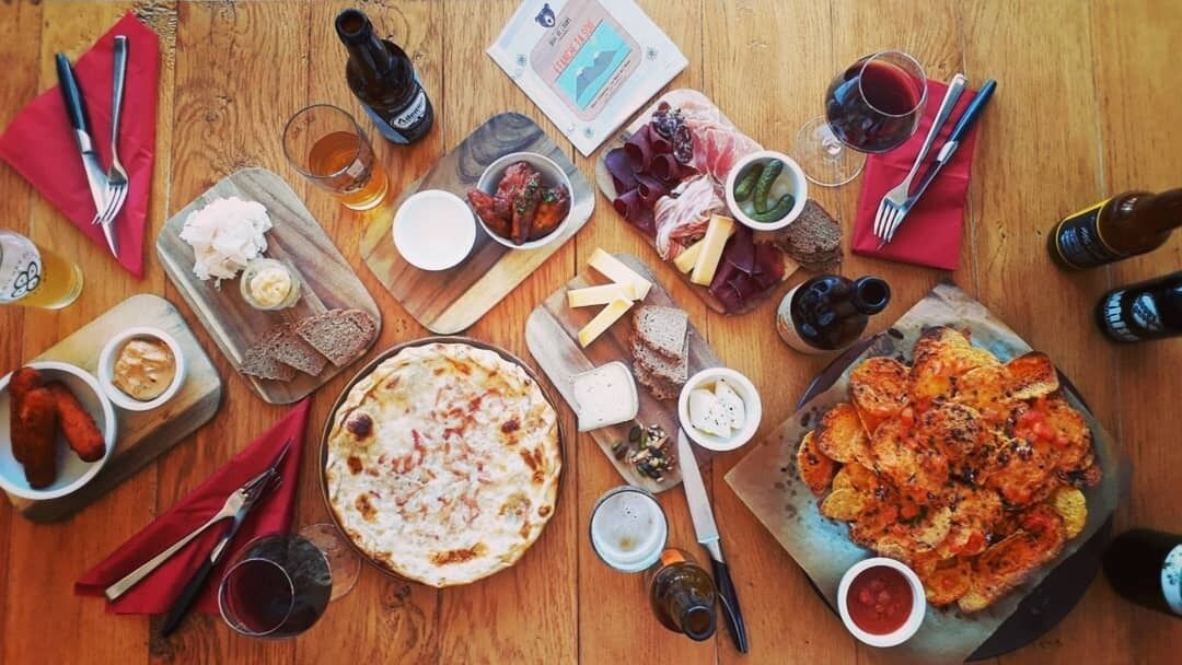Una mesa de madera con diversos alimentos, como pizza, embutidos y pan, junto con vasos de cerveza y botellas.