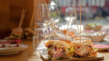Un ambiente de restaurante con sándwiches sobre tablas de madera, vasos y un fondo borroso de otros alimentos.