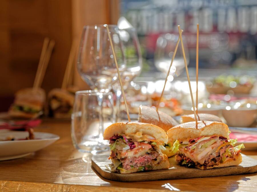 Un ambiente de restaurante con sándwiches sobre tablas de madera, vasos y un fondo borroso de otros alimentos.